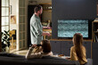 © pressmaster - Caucasian man standing near television with static screen gesturing while two Caucasian wife and child sitting on sofa holding remote control watching television together