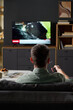 © pressmaster - Caucasian man sitting on sofa watching television holding remote control in living room, back view showing short hair and relaxed posture, news program on screen
