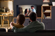 © pressmaster - Caucasian young adult couple sitting on sofa watching television together, man holding remote control, both facing screen, spending leisure time in modern living room interior