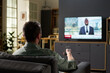 © pressmaster - Caucasian man sitting on sofa holding remote control watching television news broadcast featuring Black man in suit standing in modern living room interior