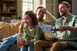 © pressmaster - Caucasian girl smiling while sitting on sofa with Caucasian woman and man, man holding remote control and bowl of popcorn, girl holding drink with straw, family watching television together