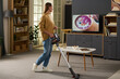 © pressmaster - Caucasian young adult woman vacuuming carpet in modern living room while watching television, standing near coffee table with remote control and bowl of snacks visible on surface