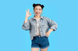 © STOCK88PHOTOGRAPHER - young Asian woman winking and making an okay gesture against a vibrant backdrop. She looks stylish and approachable on a blue background