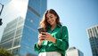 © miss irine - Young woman smiles looking at phone outdoors near modern glass buildings. She wears a green shirt. Business woman uses smartphone in city. Clear blue sky.