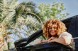 © SHOTPRIME STUDIO - Smiling woman with curly hair leaning out car window, sunny day with palm trees in background, casual white blouse, happy expression outdoors.