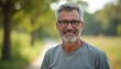 © Vadym - Mature man with grey hair, beard wears glasses smiles warmly. Stands outdoors with blurred green trees, sunny field background. Casual attire, confident look, relaxed vibe, friendly expression.