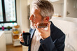 © Marko Geber - Mature businessman using smartphone speakerphone in home kitchen