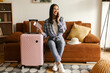 © Home-stock - Happy woman tourist sitting on sofa with passport and packed suitcase nearby, talking on phone at home, calling taxi, copy space