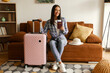 © Home-stock - Happy woman preparing for trip while sitting on couch with packed suitcase and passport in hand, smiling at camera