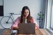 © Daniel - A happy young woman sits at a desk at home, smiling while browsing the internet on her laptop, enjoying modern technology and a comfortable remote work lifestyle in her living room