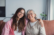 © Daniel - A smiling mature mother and her adult daughter sit on the couch at home, hugging and looking at the camera, sharing a warm and emotional moment of family connection and love