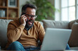 © Pete - Man in glasses shouts on cell phone while working on laptop at home. He is sitting on a couch looking stressed and frustrated with a work issue. His facial expression is angry.