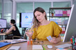 © H_Ko - Focused woman in a yellow shirt holds a glass of water and medication while working at her desk in a modern office. A busy colleague is seen in the background.