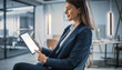 © Zainu - A professional woman sitting in a modern office environment looking at a digital tablet with a thoughtful expression from a slightly angled viewpoint.