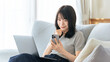 © Trickster* - 自宅のソファでスマートフォンを確認しながらノートパソコンを使う女性,在宅ワークの日常, A Woman Checking Her Smartphone While Using a Laptop on a Sofa at Home
