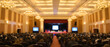 © itlada - Conference hall filled with audience at seminar featuring an illuminated stage with presentation screens under golden chandeliers in a blurred corporate event background