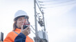 © Happy Photo - A worker stands near power lines, holding a radio in hand. The worker talks into the device while wearing a hard hat and bright safety clothes