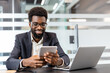 © Liubomir - African american businessman smiling, connecting with a digital tablet while working in a bright modern office, showcasing professional expertise, communication, and innovative technology