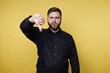 © bodnarphoto - Man showing thumbs down gesture in front of yellow wall during indoor scene