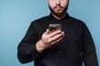 © bodnarphoto - Man in black shirt holds smartphone in front of blue background while looking at the screen and standing still