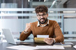 © Liubomir - Young man smiling and clenching his fist in triumph while holding an envelope with important news, celebrating a career achievement or promotion in a modern office workplace