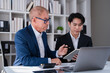 © Tj - Experienced senior businessman providing mentorship and guidance to a young male colleague, discussing business strategy and financial data during a corporate meeting in a modern office environment