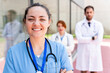 © StockMe - Smiling female nurse looking at camera, standing with arms crossed, male and female doctors blurred in background