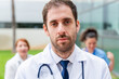 © StockMe - Doctor looking seriously at camera, medical team standing behind him outdoors, healthcare occupation