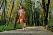 © SHOTPRIME STUDIO - Woman running in bamboo forest path, smiling and wearing orange skirt and sneakers, outdoor jog for fitness and travel, healthy young adult enjoying nature and sunlight on gravel trail