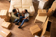 © DC Studio - Young black couple sits on floor of new apartment surrounded by moving boxes, talking and planning decor. Spouses share conversation in unfurnished living room while unpacking belongings together.