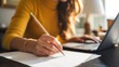 © Karuna - Creative Writing Moment: A focused individual engrossed in creative writing, meticulously crafting words on paper with a pencil while surrounded by a laptop, emphasizing the art of composition.