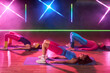© pressmaster - Three young adult women exercising on yoga mats performing glute bridge pose in fitness studio with neon lights, engaging in group workout session