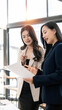 © NAMPIX - Two professional Asian businesswomen standing and discussing business documents in a bright, modern office.