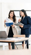 © NAMPIX - Two professional Asian businesswomen standing and reviewing financial reports on a tablet and clipboard in a bright modern office workspace.