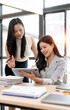 © NAMPIX - Two professional Asian businesswomen smiling while discussing data on a digital tablet in a bright modern office.