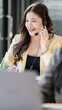 © NAMPIX - Portrait of a smiling Asian customer service representative with a headset working on a laptop in a bright modern office.