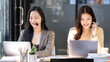 © NAMPIX - Two smiling Asian customer service agents wearing headsets and working on laptops in a modern office call center.