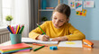 © Paradoxx - A young girl in a yellow shirt drawing with colored pencils on paper at a wooden desk with school supplies in a classroom setting.