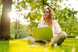 © maxbelchenko - A female freelancer sits in a green meadow with a laptop on a sunny day. A cheerful woman wearing glasses works on a laptop in park at sunset. Concepts of remote work, relaxation, the outdoors.