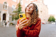 © maxbelchenko - A cheerful woman with wireless headphones and a phone enjoys the urban environment on a sunny day. A young woman listens to music and strolls through the old town. Concept of rest and relaxation.