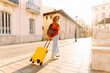 © maxbelchenko - A beautiful traveler with a yellow suitcase and backpack walks along a city street on a sunny day. The young woman in sunglasses and a hat enjoys the stroll. Concept of tourism and leisure.