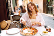 © maxbelchenko - Portrait of a beautiful woman at a restaurant table, holding a slice of fragrant pizza. A cheerful woman enjoys a delicious Italian pizza at sunset. Concepts: food, tourism, enjoyment.