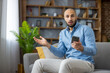 © Liubomir - Surprised man sitting on sofa, holding mobile phone, feeling disbelief and confusion while reacting to unexpected shocking news or a surprising message at home