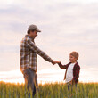 © Acronym - Farmer and his son in front of a sunset agricultural landscape. Man and a boy in a countryside field. Fatherhood, country life, farming and country lifestyle concept.