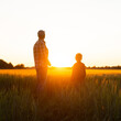 © Acronym - Farmer and his son in front of a sunset agricultural landscape. Man and a boy in a countryside field. Fatherhood, country life, farming and country lifestyle concept.