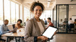 © Monktwins - Smiling professional woman holding tablet with blank screen in modern coworking office. Business presentation mockup, diverse team in background, digital device mockup for UX/UI and corporate use