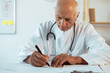 © Andrii Lysenko - Senior elderly male doctor or medical professional wearing white uniform with stethoscope working at desk healthcare in his office workplace. Medicine, healthcare and medical