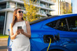 © vladim_ka - Smilling woman in a white dress uses a smartphone while charging an electric vehicle outdoors