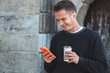 © tetxu - Young man checking his phone while drinking a takeaway in the historic area of ​​the city