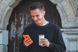© tetxu - Young man checking his phone while drinking a takeaway in the historic area of ​​the city